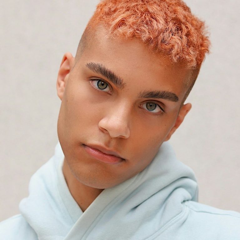 Young person with short orange afro hair, wearing a light blue hoodie, looking directly at the camera.